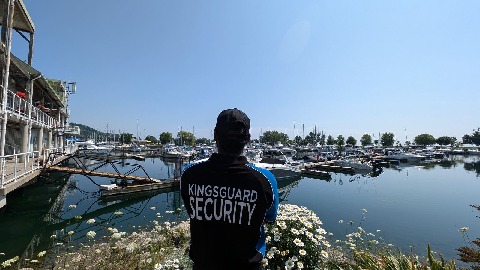 A KingsGuard guard in a branded shirt and cap overlooking a marina full of moored boats on a clear summer day