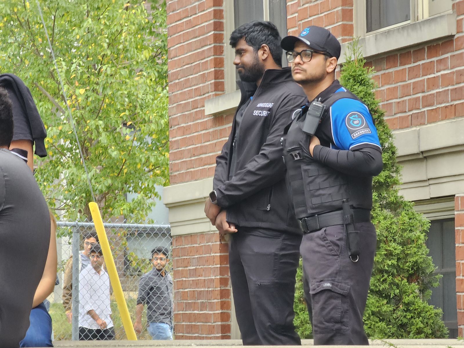 Two KingsGuard guards standing watch beside a brick campus building during a busy student gathering