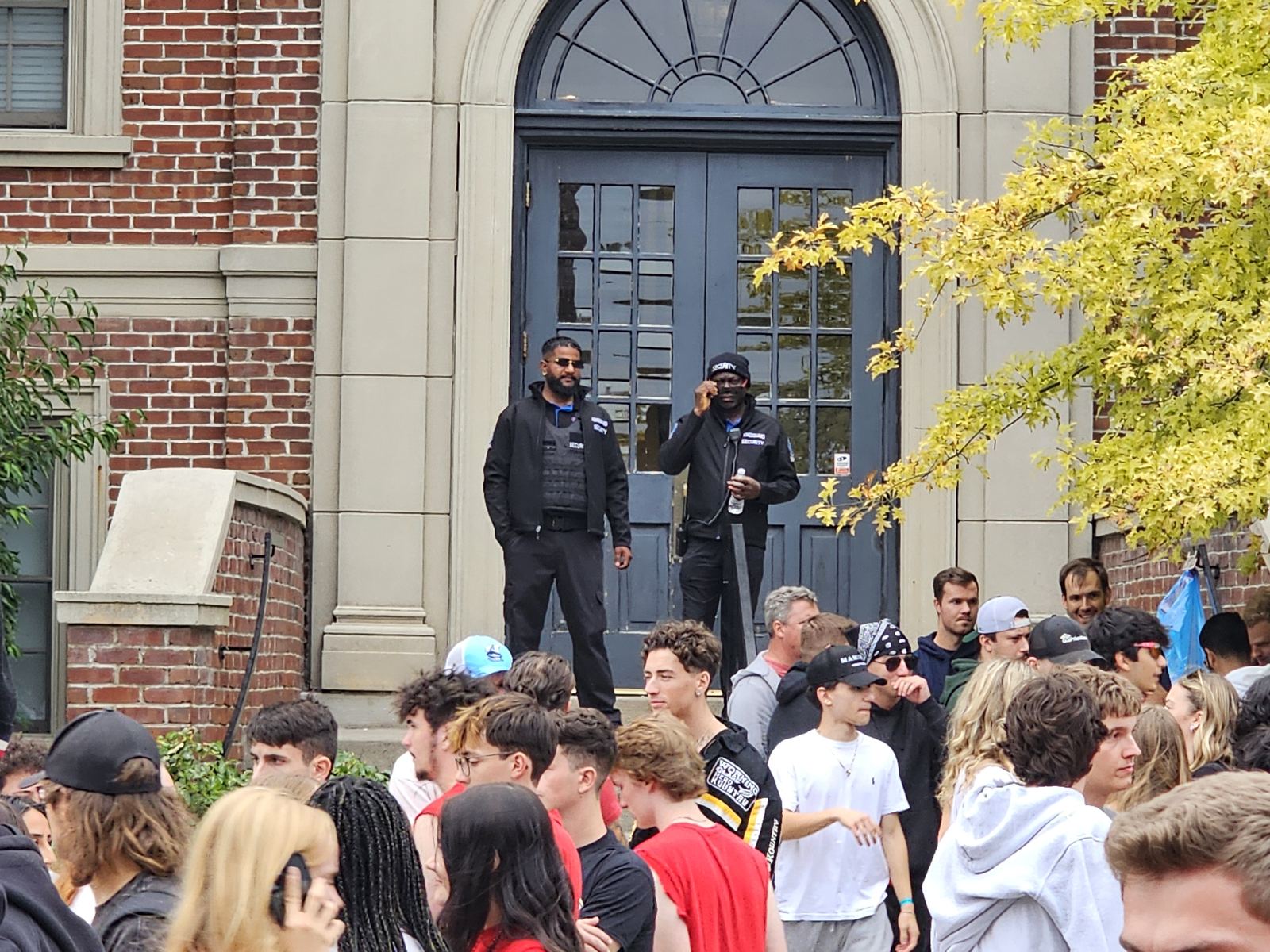 Two KingsGuard guards posted on the steps of a brick building above a large outdoor crowd at a community event