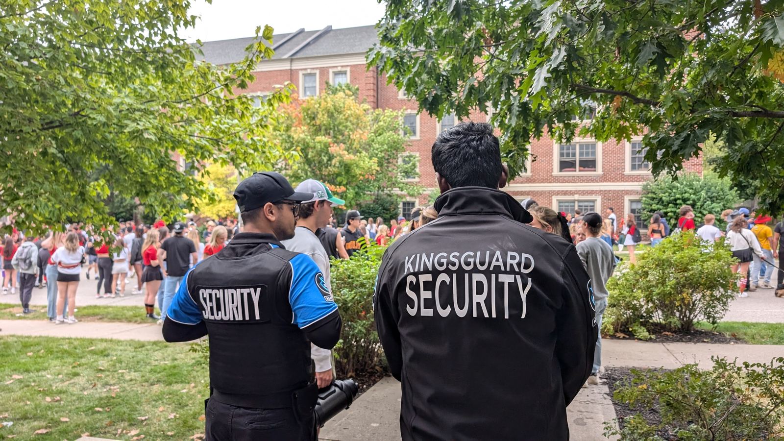 Two KingsGuard guards in branded jackets watching a large student crowd outside a campus building