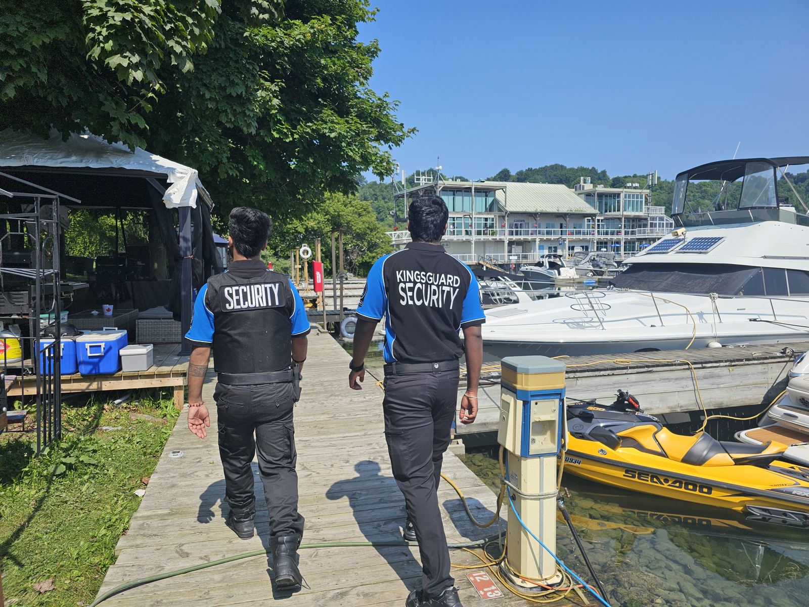 Two KingsGuard guards walking a wooden marina dock past moored boats on a mobile patrol