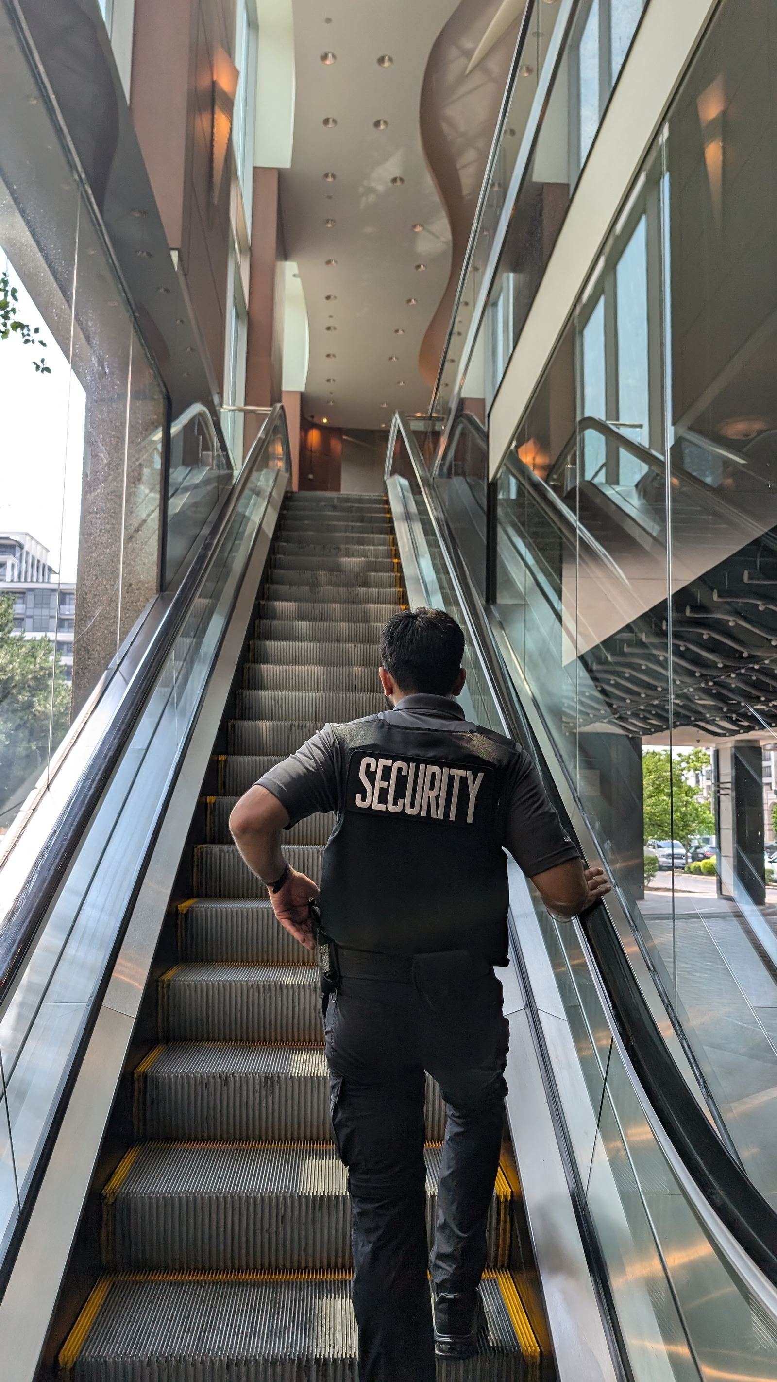 A KingsGuard guard in a marked security vest walking up the escalator of a commercial office lobby on patrol